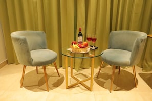 Duas poltronas azul-claras voltadas uma para a outra, com uma mesa de vidro entre elas, uma garrafa de vinho e uma cesta de frutas sobre a mesa.