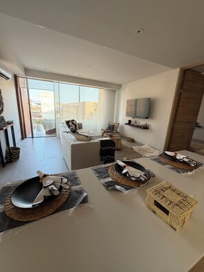 Een moderne woonkamer met een bank, een eettafel gedekt met placemats en een middelpunt, een aan de muur bevestigde televisie en een schuifpui naar een buitenruimte.