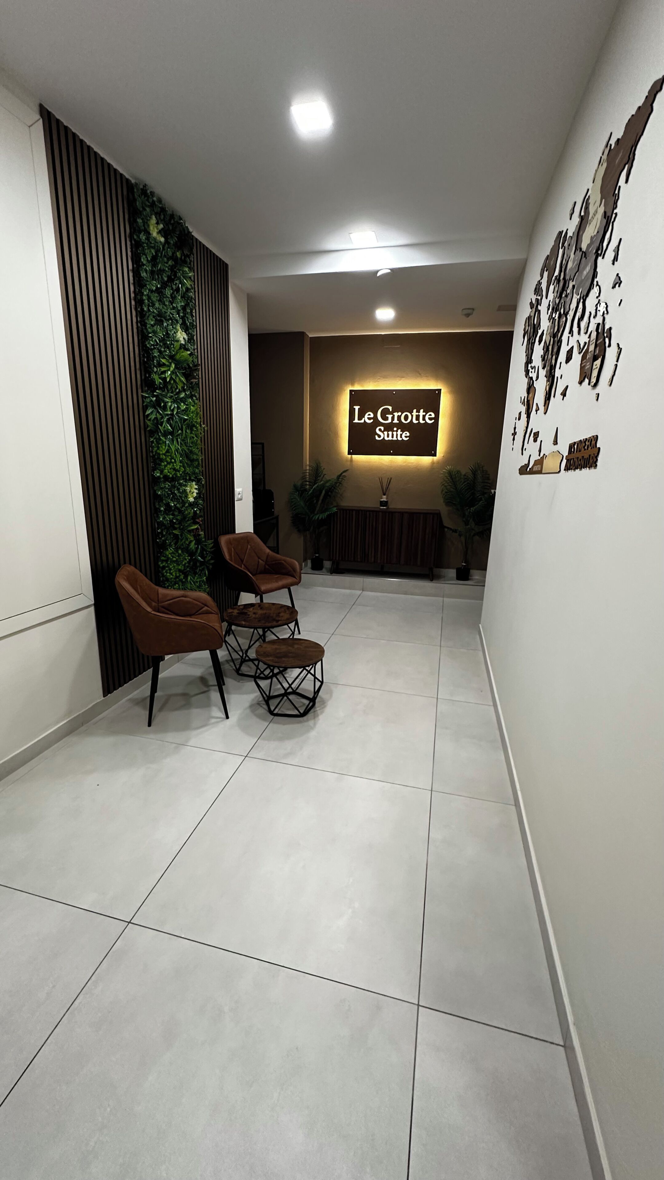 Un hall d'hôtel avec une enseigne indiquant « Le Grotte Suite », une plante fixée au mur, deux fauteuils marron et un sol carrelé.