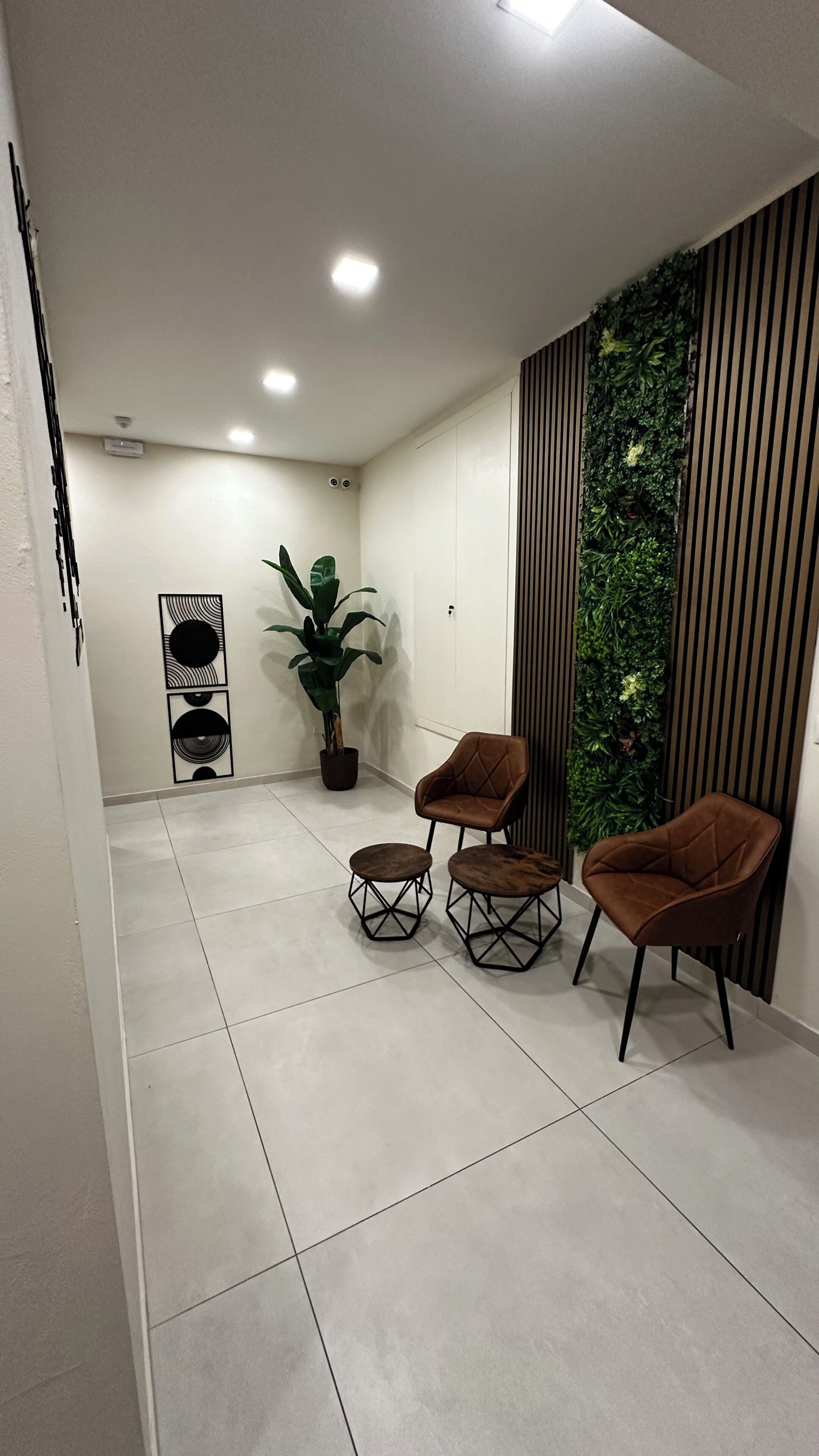 Un intérieur moderne avec un coin salon comprenant deux fauteuils marron et deux petites tables rondes, une plante en pot et un mur agrémenté de stores verticaux et de plantes grimpantes.
