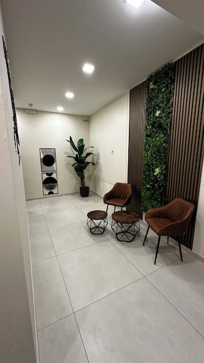 A modern interior with a seating area featuring two brown chairs and two small round tables, a potted plant, and a wall with vertical blinds and greenery. - Le Grotte Suite  (Naples)