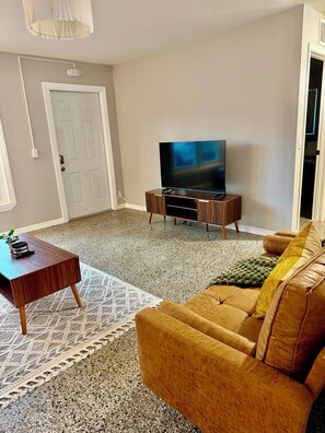 Ein Wohnzimmer mit einem Flachbildfernseher auf einem Holzständer, einem braunen Sofa, einem Couchtisch und einem gemusterten Teppich.