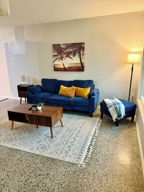 Un salon avec un canapé bleu, une table basse en bois, un petit banc et un tableau représentant une scène de plage.