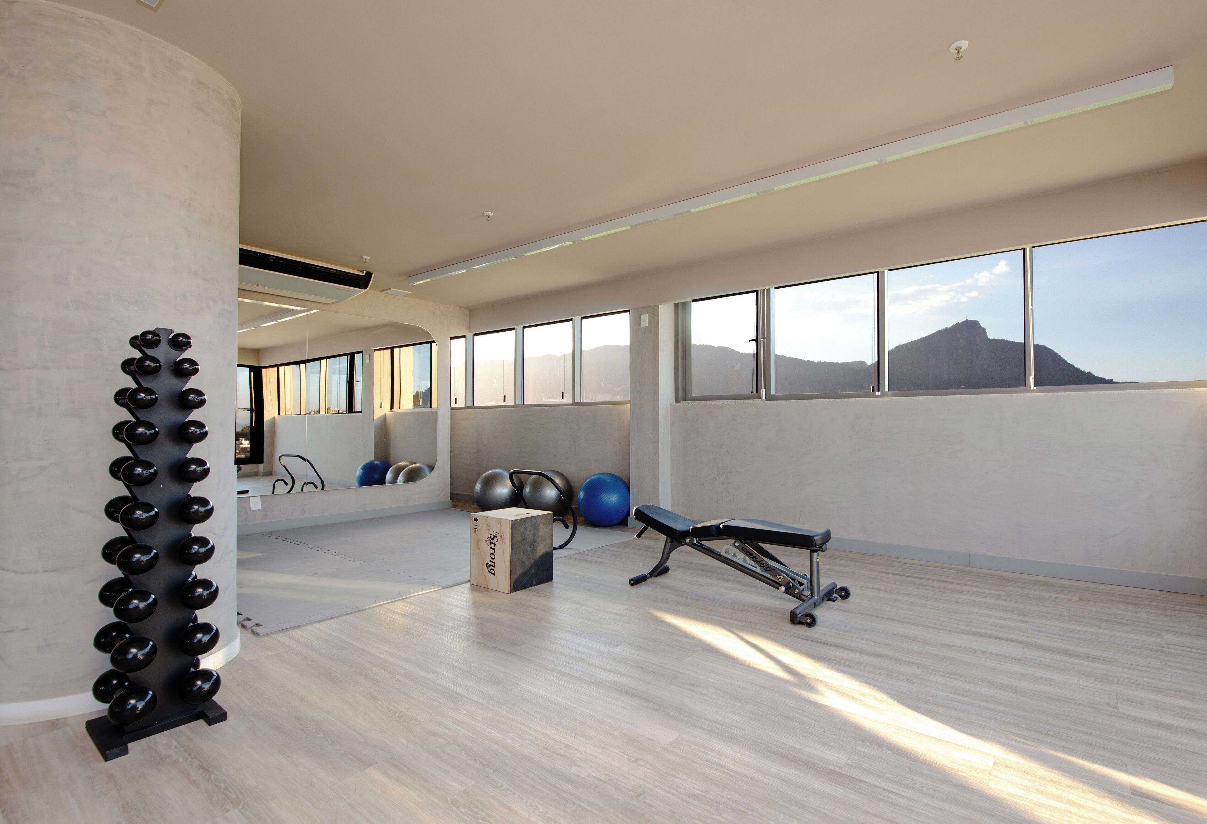 Un gimnasio amplio con equipo de ejercicio, ventanas grandes y vistas a una colina.