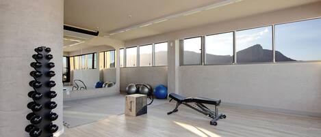 Un gimnasio amplio con equipo de ejercicio, ventanas grandes y vistas a una colina.