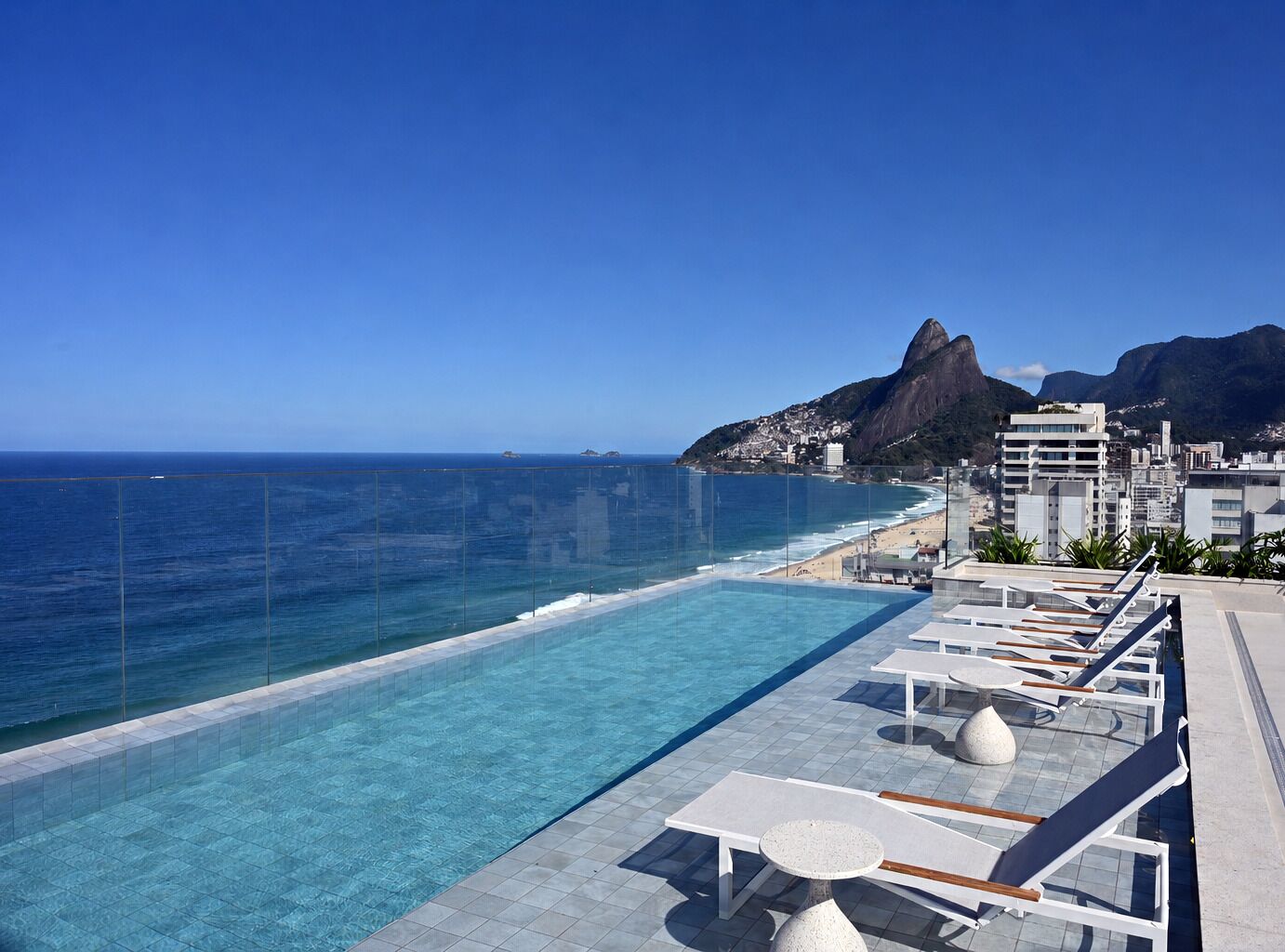 A clear blue sky, a swimming pool with lounge chairs, and a view of the ocean and mountains.