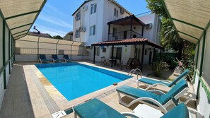 Ein Schwimmbecken mit Liegestühlen, eine überdachte Terrasse und ein zweigeschossiges Gebäude mit Balkonen.