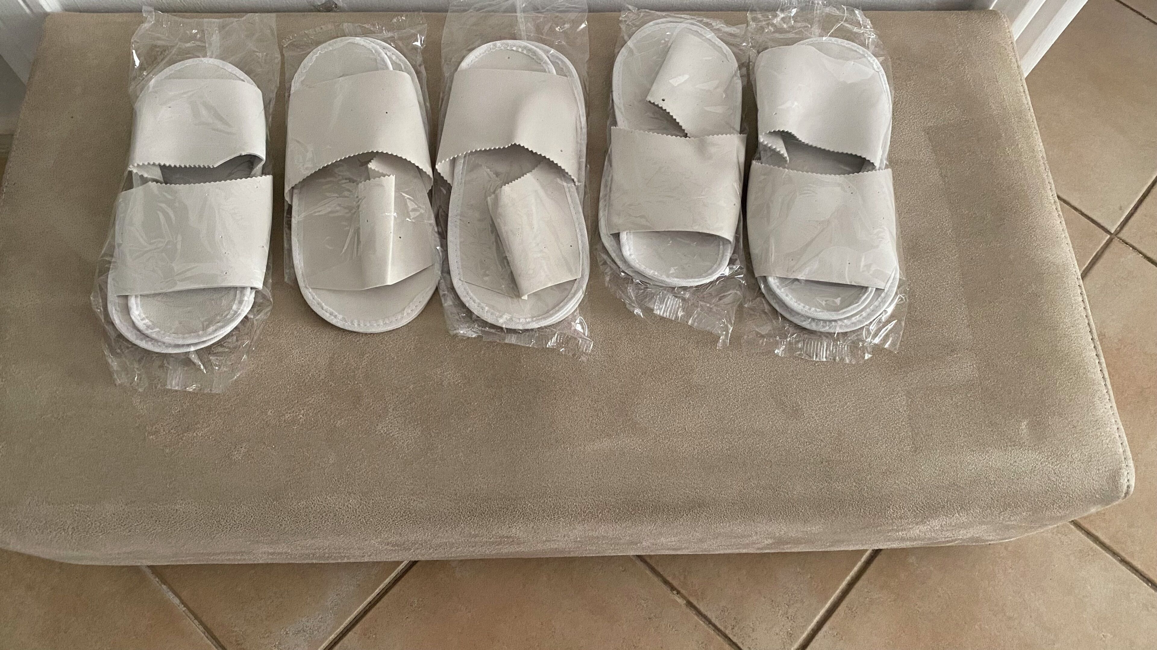 A set of five white slippers wrapped in plastic, placed on a beige cushioned surface.