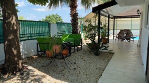 Eine überdachte Terrasse im Freien mit Tisch und Stühlen, umgeben von Blumentöpfen und einer Palme.