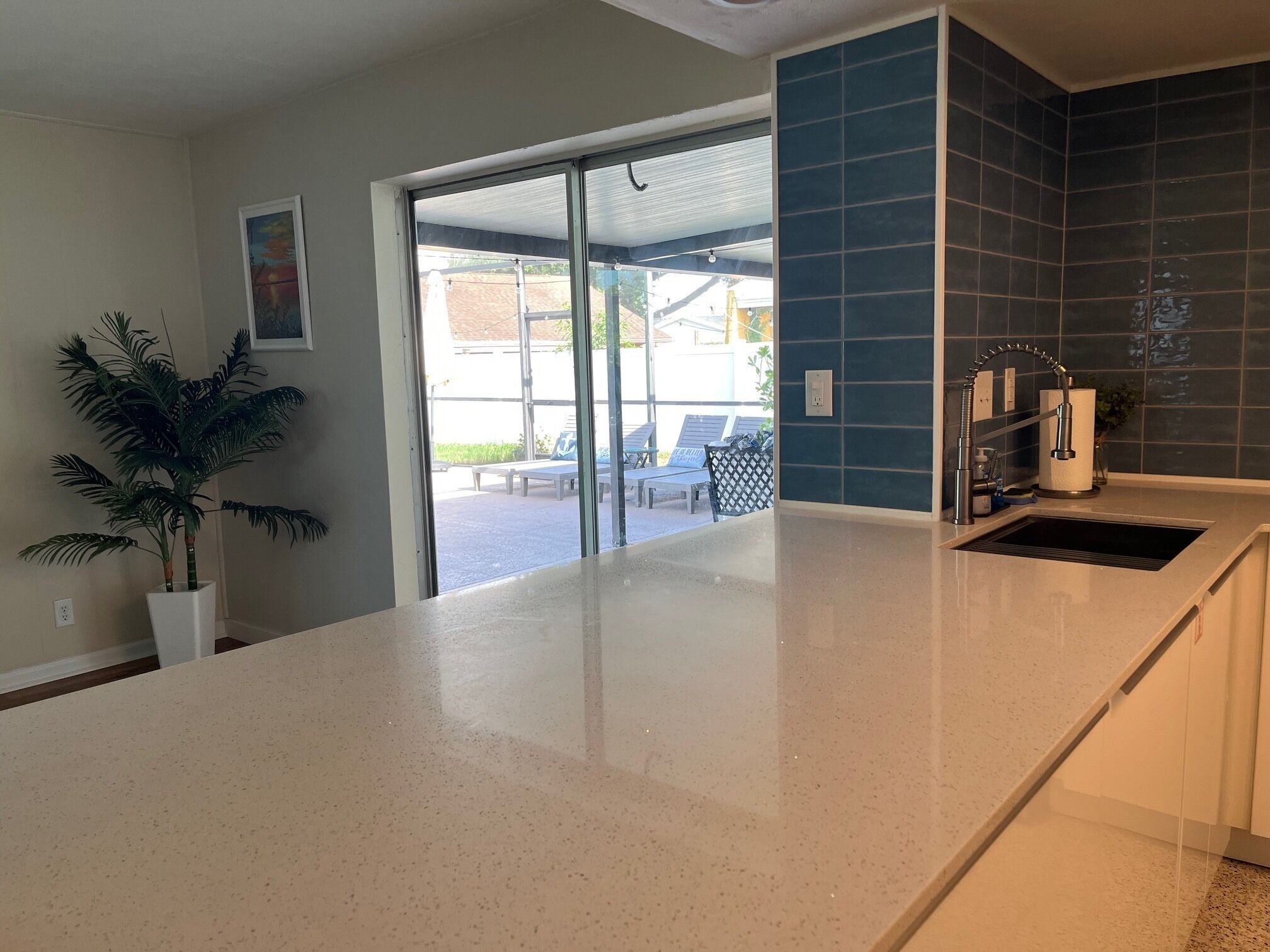 A kitchen with a white countertop, a sink, and a blue backsplash. There is a potted plant on the left and a view of outdoor seating through a glass door.