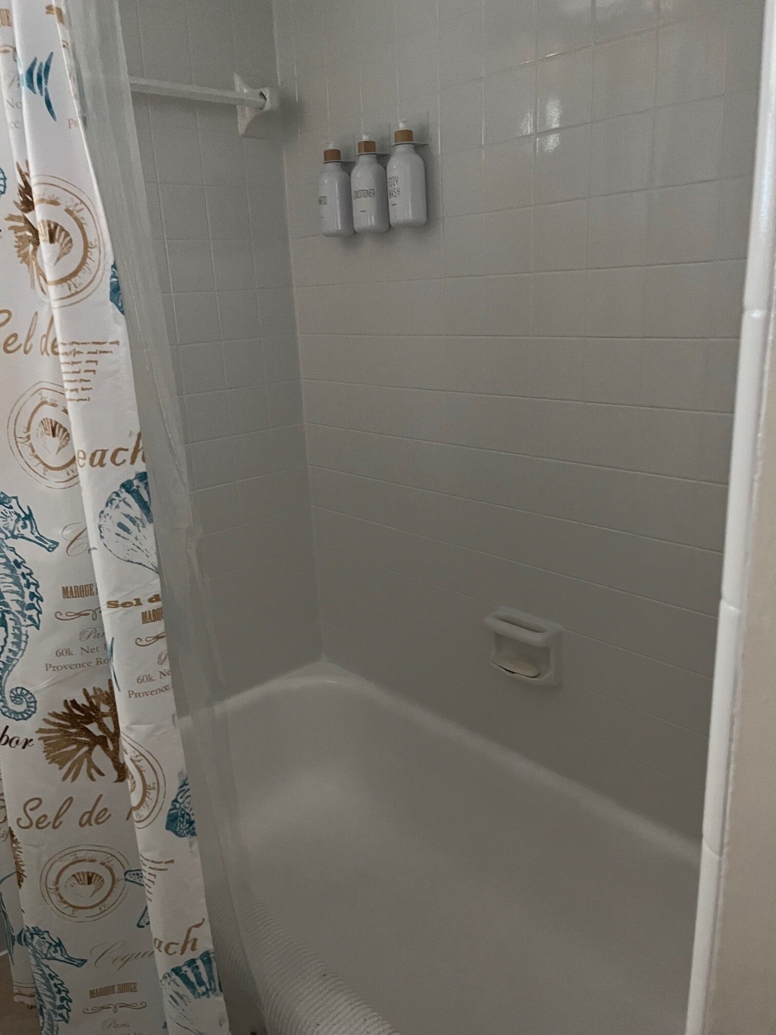 Un espace douche avec un mur recouvert de carreaux blancs, un rideau de douche orné de motifs représentant des hippocampes et trois bouteilles posées sur le mur.