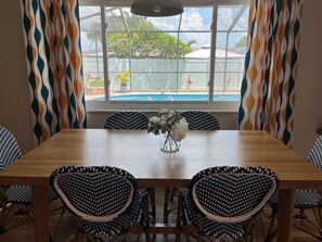 A dining area with a wooden table, black and white patterned chairs, a vase with white flowers, and a view of a swimming pool and flamingos through a window with patterned curtains. - 3-bedroom family friendly with pool in wonderful Largo just minutes to beaches (Largo)