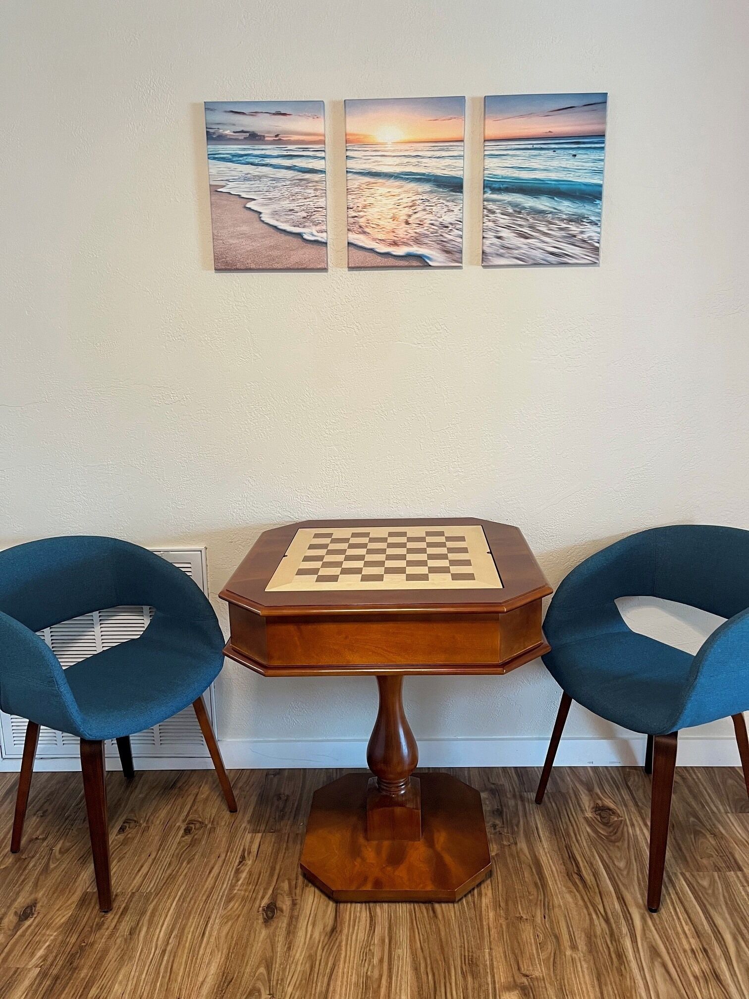 A wooden table with a chessboard in the center, flanked by two blue chairs, set against a wooden floor with a white wall in the background.