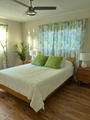 Ein Schlafzimmer mit einem Bett, einem Deckenventilator, einem Nachttisch mit Lampe und einem Fenster mit Vorhängen.
