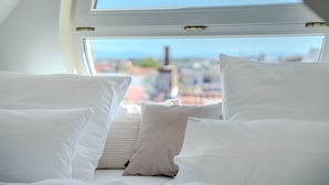 Cama com roupa de cama branca e almofadas listradas, janela de claraboia e vista para a paisagem urbana.