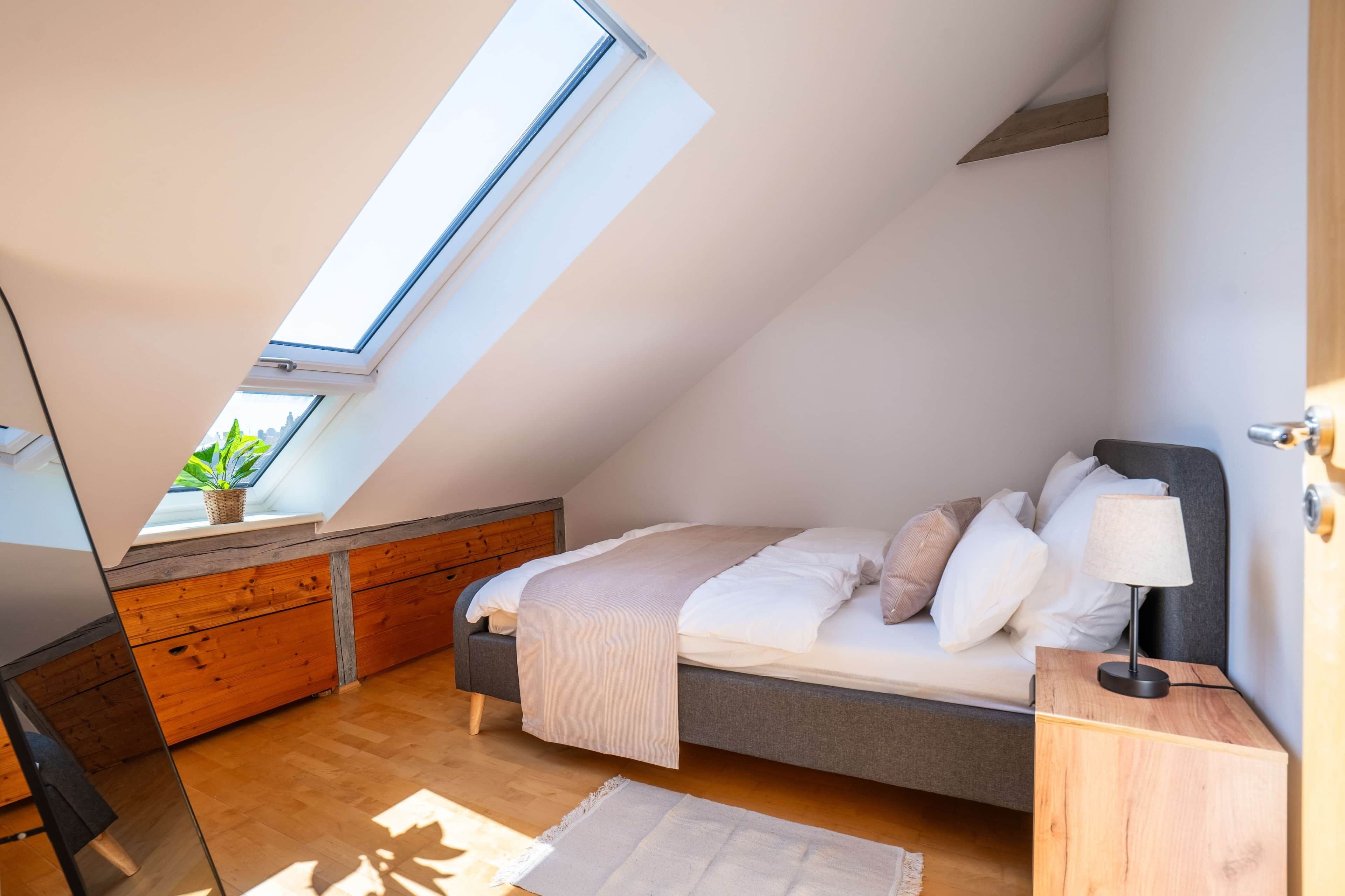 Une chambre avec un plafond en pente, un puits de lumière, un lit avec des oreillers, une table de chevet et une lampe.