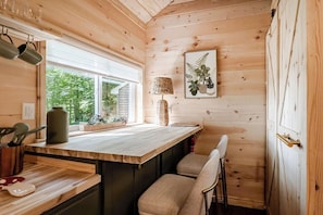 A wooden cabin interior with a kitchen area, a dining table, and a window overlooking greenery.