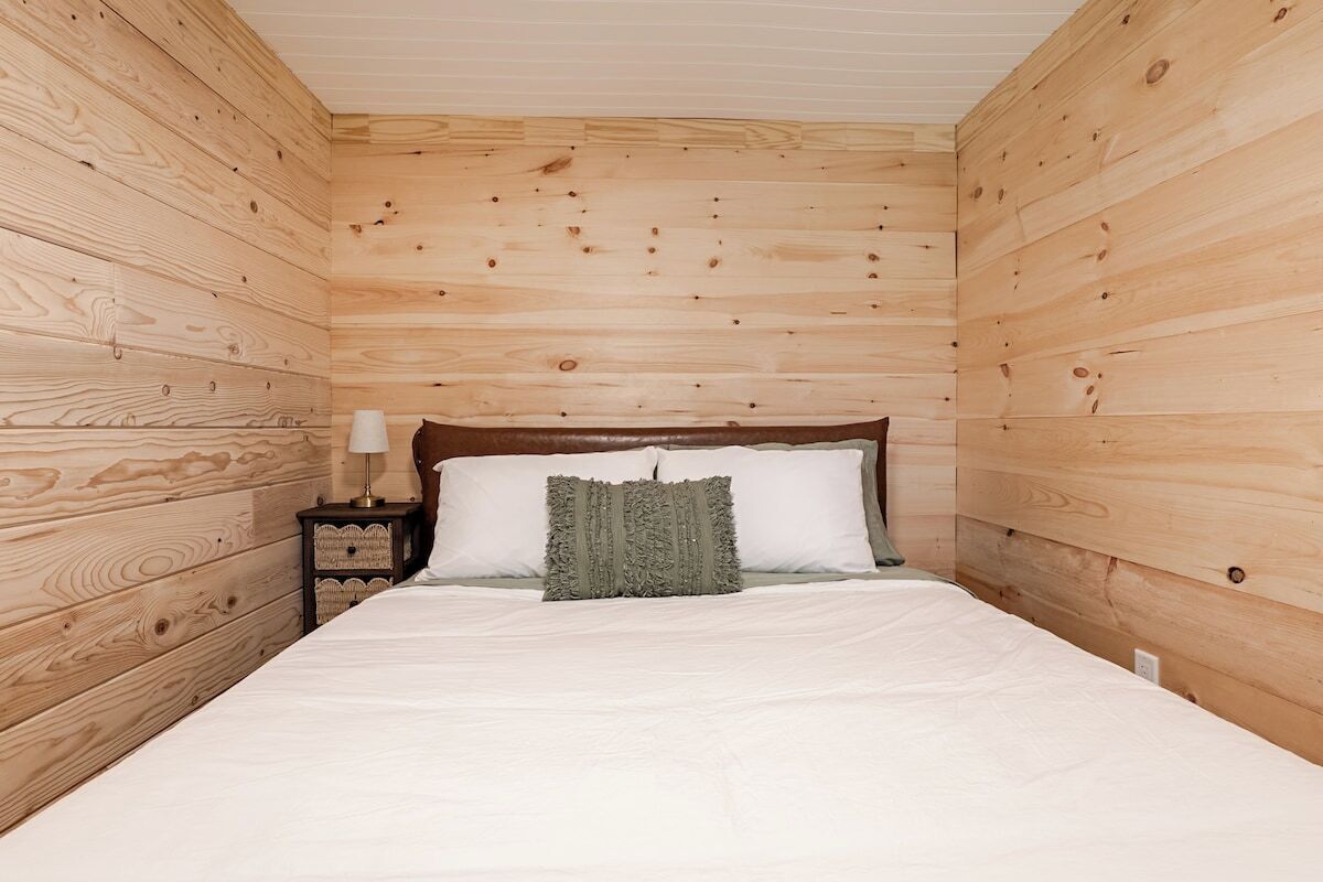 Une pièce avec un mur lambrissé en bois, un lit avec une literie blanche, une table de chevet avec une lampe et un coussin décoratif en tissu tissé.