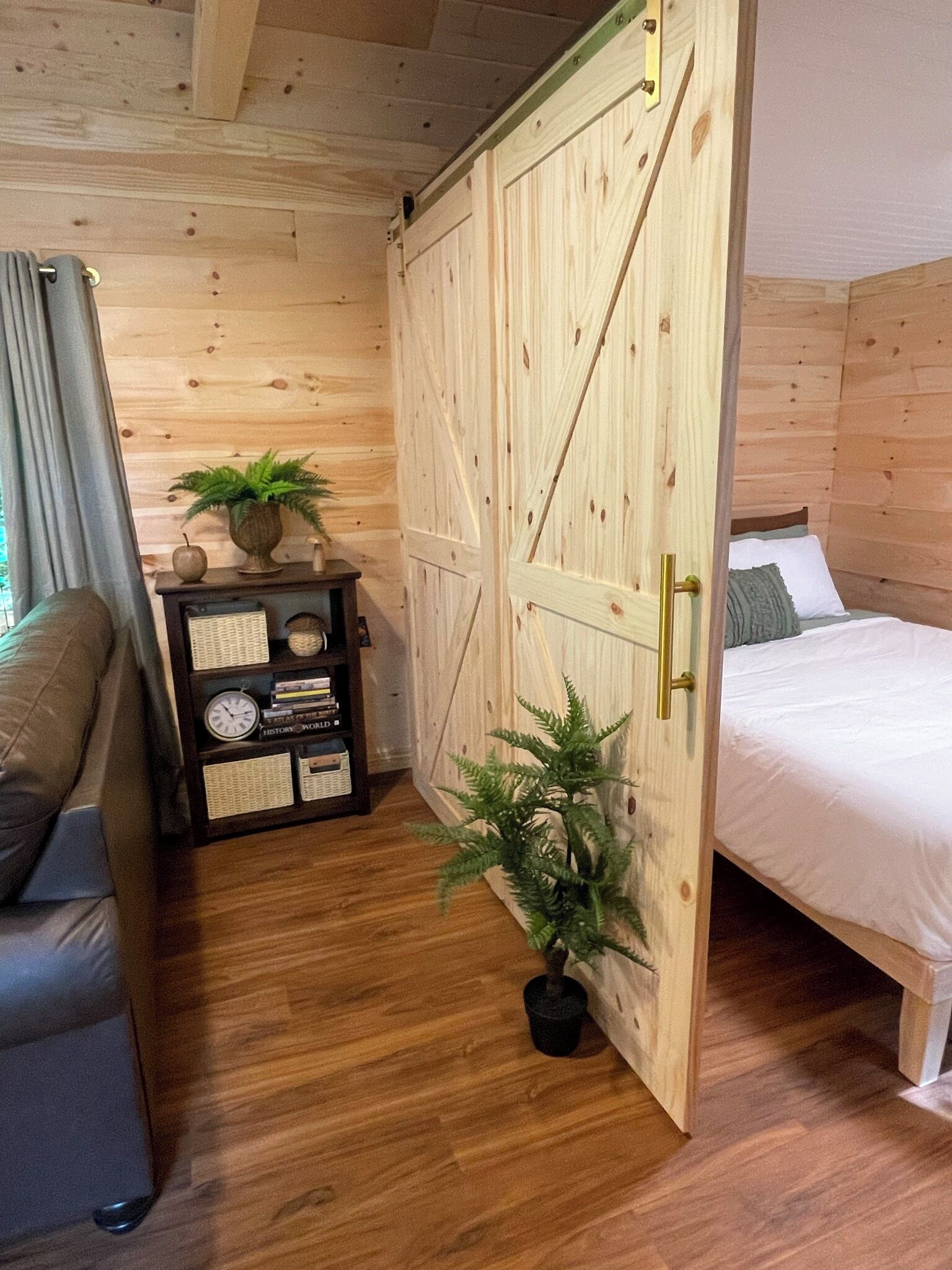 A wooden room with a bed, a small table with a clock, and a potted plant.