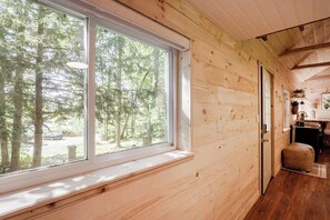 Un intérieur en bois avec une grande fenêtre donnant sur une forêt, une porte menant à la cuisine et un banc placé près de la fenêtre.