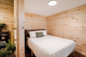 Une chambre avec un mur en bois, un lit avec une literie blanche et une petite plante posée sur une étagère.