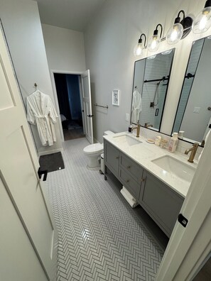 Un bagno moderno con pavimento a spina di pesce, un mobile lavabo bianco con un grande specchio e una zona doccia visibile tramite una porta in vetro.