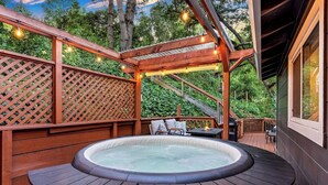 Ein Whirlpool, umgeben von Holzterrasse-Möbeln, einer Holz-Pergola mit Lichterketten und Blick auf Bäume.