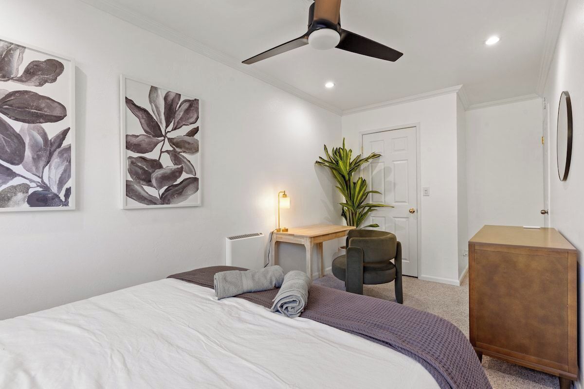 Une chambre à coucher avec un lit, un bureau, une chaise, une plante et un ventilateur de plafond.