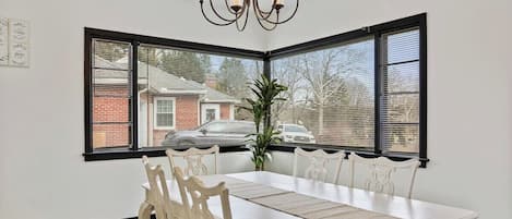 A dining area with a white table, matching chairs, a chandelier, and a view through large windows.