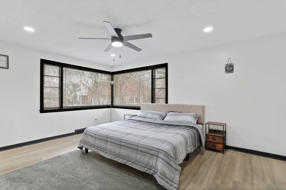 Une chambre à coucher comprenant un lit, une table de chevet, une commode, un ventilateur de plafond et une fenêtre avec des stores.