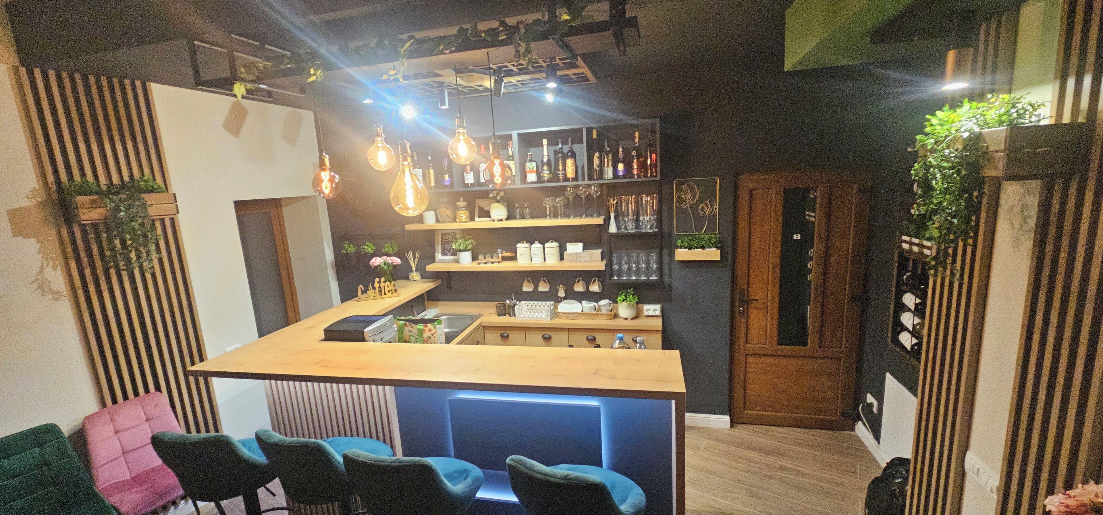 Una zona de bar con mostrador de madera, asientos y estanterías con botellas y vasos.