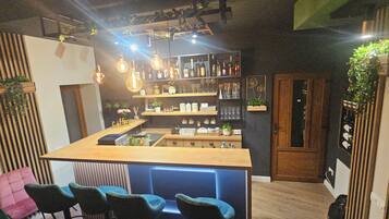 Una zona de bar con mostrador de madera, asientos y estanterías con botellas y vasos.