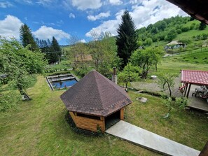 A wooden cabin with a sloped roof, surrounded by a grassy yard, a small pond, and a red-roofed structure.