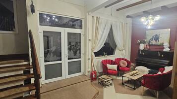 A living room with a piano, red chairs, a wooden staircase, and a window with curtains.