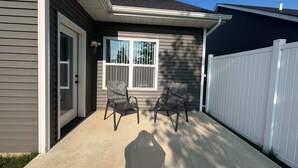 A house with a covered patio, two chairs, a white fence, and a window with blinds.
