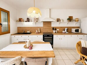 A modern kitchen with white cabinetry, a wooden countertop, a dining table with a white tablecloth, and a wicker chair. - Le Cocon | La Séguinière | Near Puy du Fou | 2-bedroom apartment (La Séguinière)