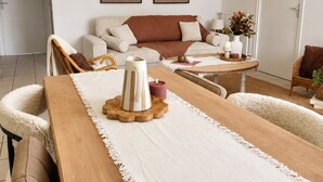 Un saló modern amb una taula de dinar de fusta, butaques blanques, un sofà i una taula de centre. Hi ha dues obres d'art abstractes penjades a la paret, una làmpada penjant de vimetlla i una porta blanca al fons.