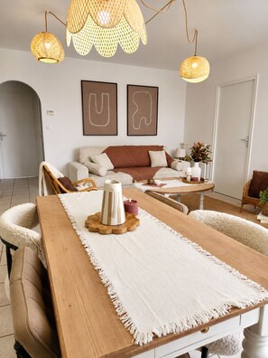 A modern living room with a wooden dining table, white armchairs, a sofa, and a coffee table. There are two wall-mounted abstract art pieces, a wicker pendant light, and a white door in the background. - Le Cocon | La Séguinière | Near Puy du Fou | 2-bedroom apartment (La Séguinière)