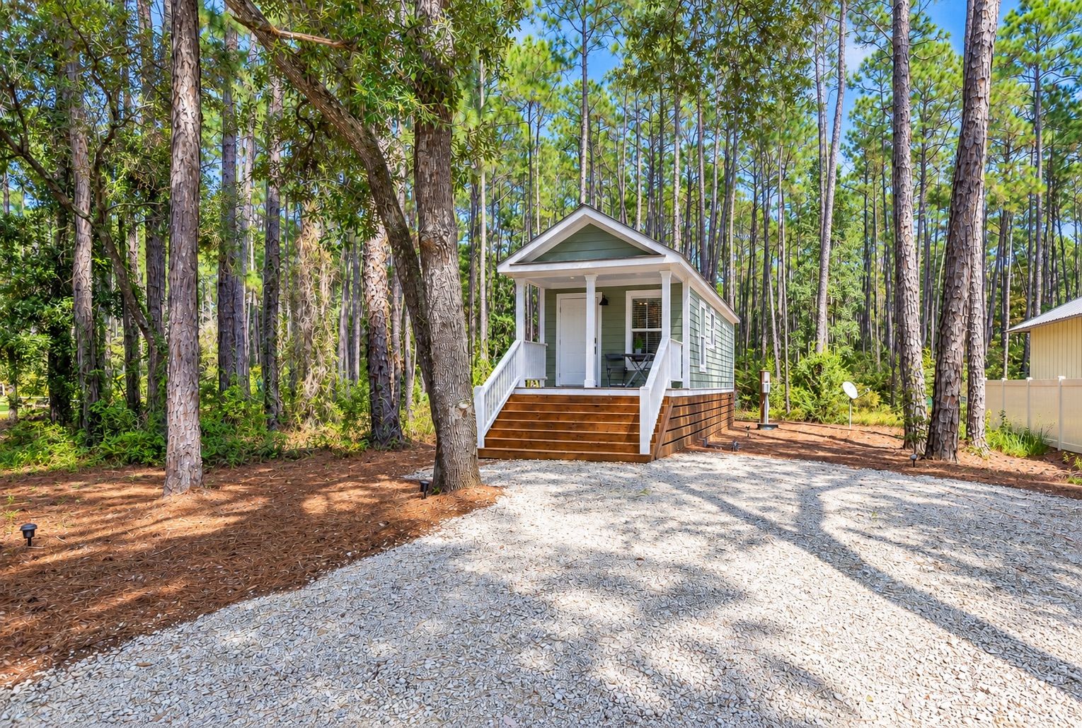A small cabin with a porch and steps, surrounded by tall trees and a gravel driveway.
