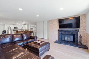 Un salon moderne avec un canapé d’angle en cuir, un téléviseur à écran plat fixé au-dessus d’une cheminée, et une cuisine en arrière-plan.