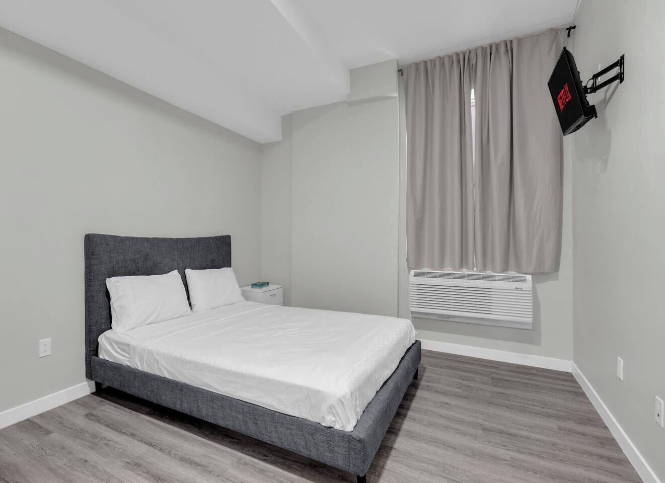 Une chambre à coucher avec un lit, une table de chevet, un téléviseur fixé au mur et une fenêtre avec des rideaux.