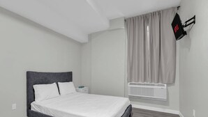 Un dormitorio con cama, mesita de noche, televisor en la pared y ventana con cortinas.