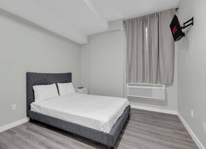 A bedroom with a bed, a nightstand, a wall-mounted TV, and a window with curtains. - The Onyx Studio • DT Oshawa (Oshawa)
