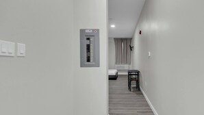 Un pasillo con un cuadro eléctrico, una cama, una silla y una ventana con cortinas.