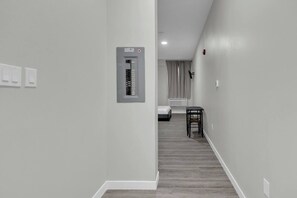 A hallway with a power panel, a bed, a chair, and a window with curtains. - The Onyx Studio • DT Oshawa (Oshawa)