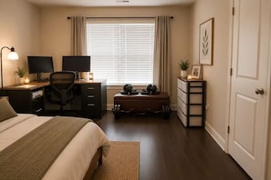 Quarto com cama, escrivaninha, dois monitores, cadeira, janela com cortinas, uma planta pequena e porta.