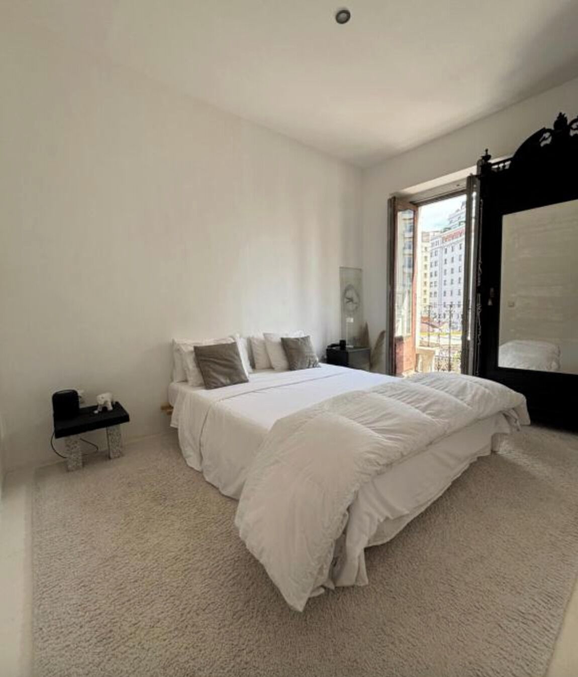 A bedroom with a large bed, a nightstand, a small table, and a window with a view of buildings.