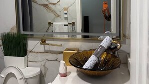 Un baño con pared de mármol, un lavamanos blanco, un espejo con detalles dorados, un inodoro y un cuenco decorativo.