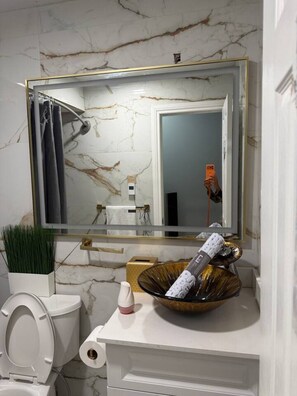 Uma casa de banho com parede de mármore, pia branca, espelho com detalhes dourados, sanita e uma tigela decorativa.