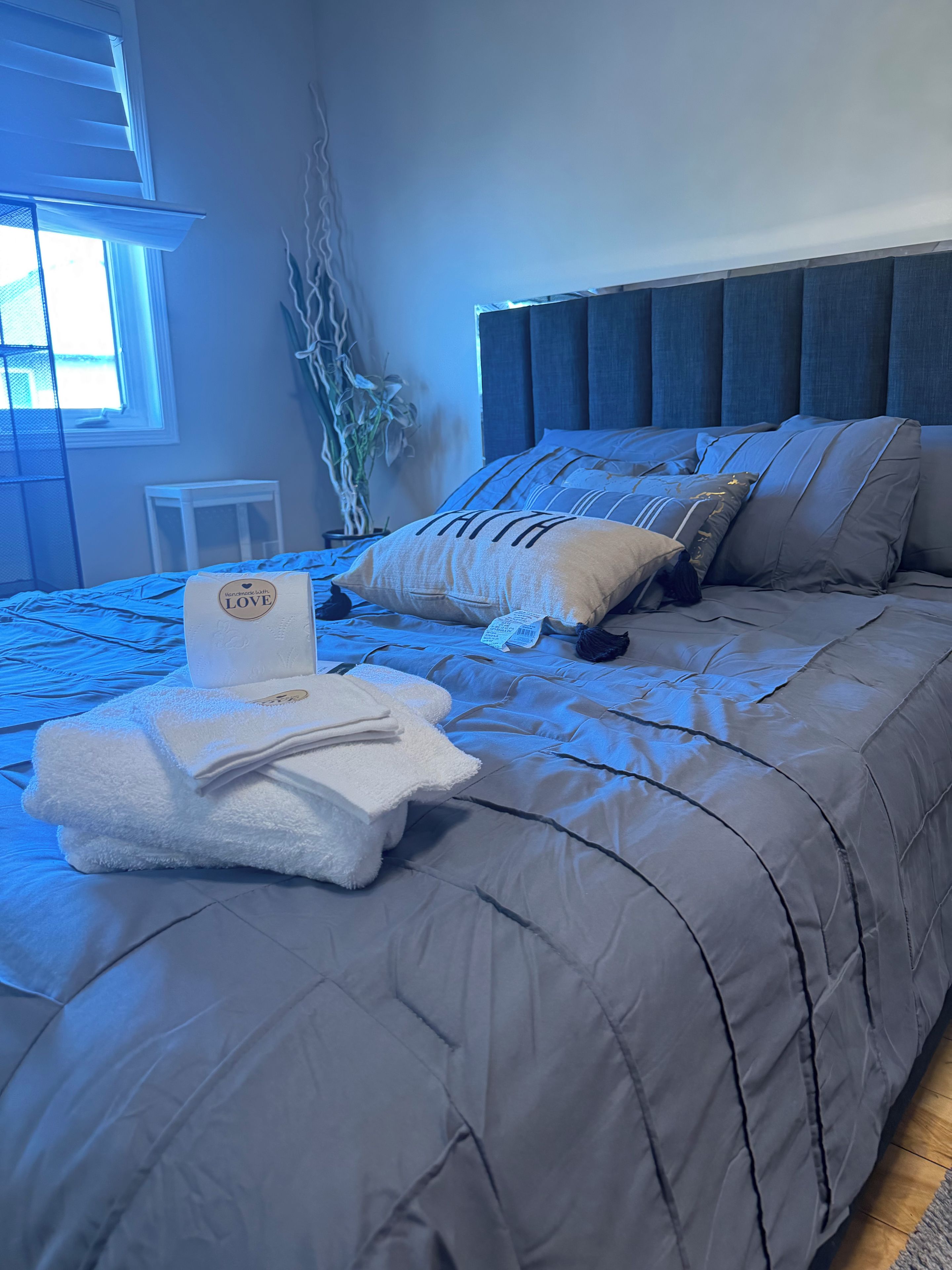 A neatly made bed with a grey duvet, a pillow with the word 'FAITH,' and a folded towel with a logo.